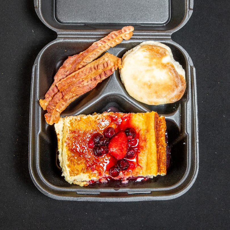 Breakfast takeout meal with crispy bacon, a biscuit, and berry-topped French toast.