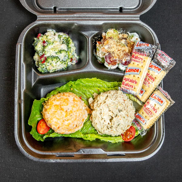 A takeout container holds broccoli, grape, pimento cheese, and chicken salads with crackers.
