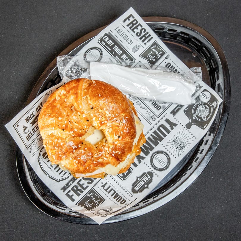 Croissant sandwich with melted cheese and sesame seeds in a basket with patterned paper and wrapped utensils.