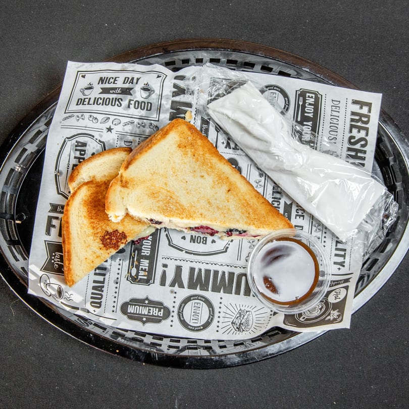 Toasted sandwich halves in a basket with patterned paper, a sauce cup, and wrapped utensils.