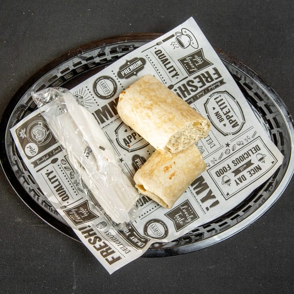 Two wraps, one revealing filling, in a basket with patterned paper and wrapped plastic cutlery.