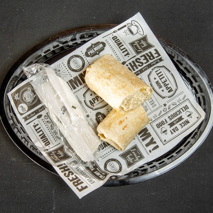 Two wraps, one revealing filling, in a basket with patterned paper and wrapped plastic cutlery.
