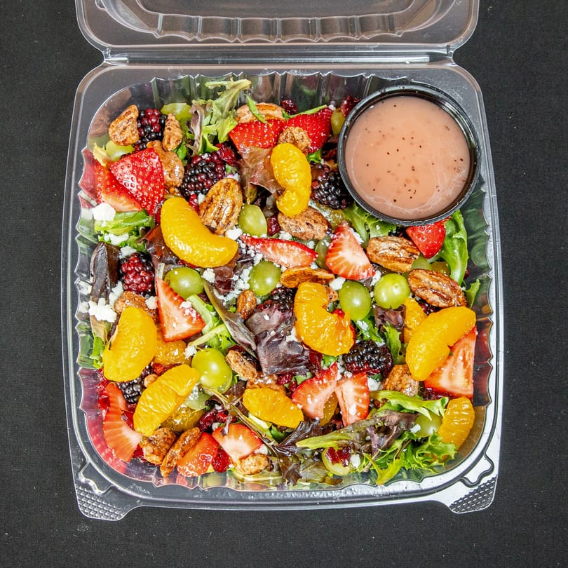 Mixed greens salad with strawberries, grapes, blackberries, oranges, pecans, feta, and dressing in a container.