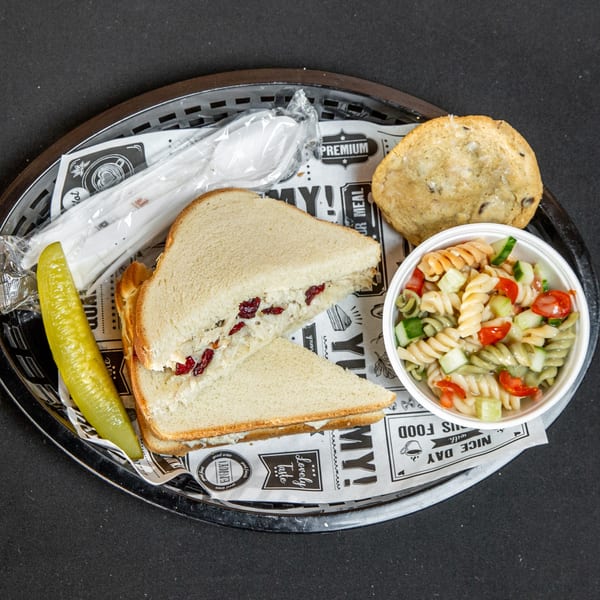 Chicken salad sandwich with cranberries, pasta salad, pickle, cookie, and cutlery in a basket.