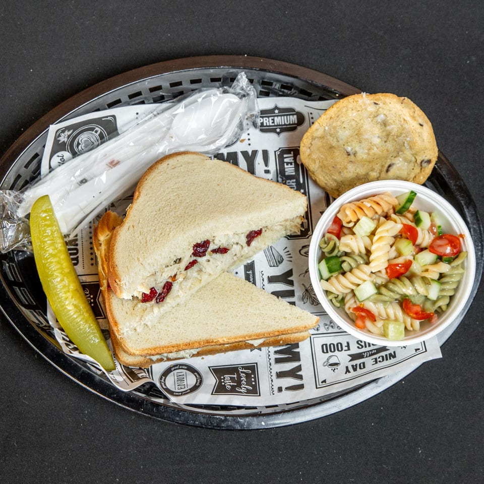 Chicken salad sandwich with cranberries, pasta salad, pickle, cookie, and cutlery in a basket.