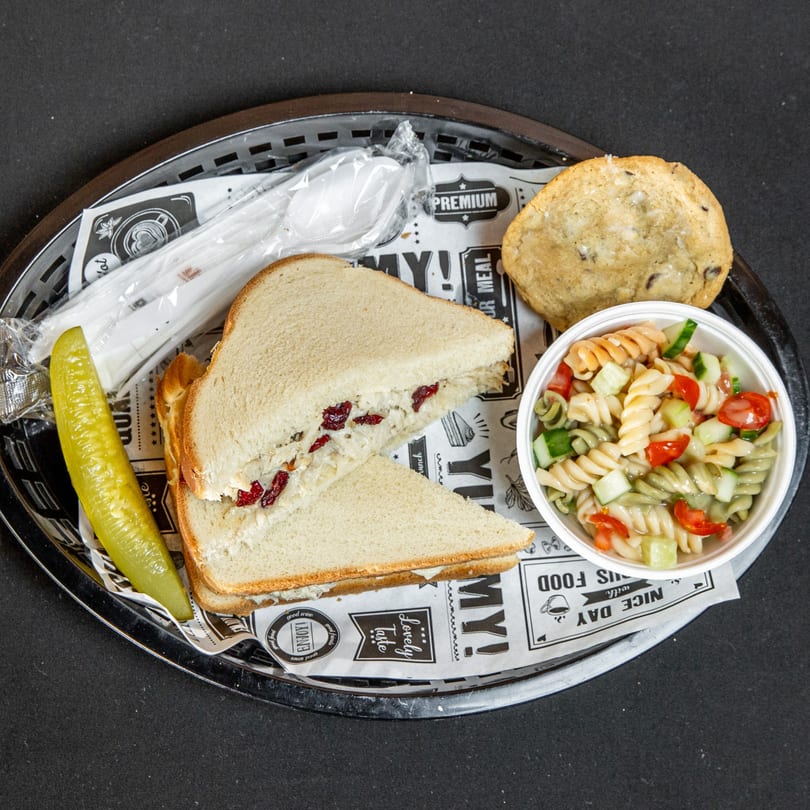 Chicken salad sandwich with cranberries, pasta salad, pickle, cookie, and cutlery in a basket.